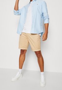 Ljusblå skjorta över en vit t-shirt, i kombination med beige uppvikta shorts och vita sneakers, mot en enkel bakgrund.