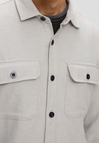 Camicia button-up grigio chiaro con tessuto testurizzato, taschini sul petto e bottoni scuri. Presenta un colletto e dettagli di cucitura visibili.