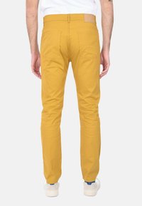 Chinos gialli in cotone con vestibilità slim, dotati di tasche posteriori e una toppa in pelle color nocciola in vita. Indossati con sneakers bianche.