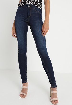 Mujer con vaqueros ajustados azul oscuro, una blusa negra con flores y sandalias de tacón beige, de pie frente a un fondo blanco liso.