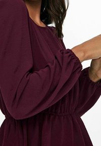 Donna che indossa un abito lungo a maniche, di colore bordeaux, con polsini e vita elasticizzati, mani sollevate vicino al petto su uno sfondo bianco.