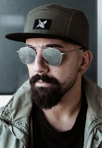 Zöld sapka fekete folttal, kerek ezüst napszemüveg, és világos olívzöld dzseki. Férfi szakálla és bajsza látható. Tiszta, modern dizájn.