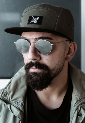 Green cap with a black patch, round silver sunglasses, and a light olive jacket. Beard and mustache visible. Clean, modern design.
