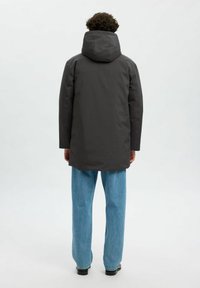 Giacca a piumino lunga, grigio scuro, con cappuccio, texture liscia, taglio dritto e tasche laterali, indossata su jeans chiari, larghi.