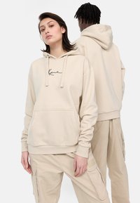 Beige hættetrøje med frontlomme og sort signaturdetalje; kombineret med matchende beige cargo-bukser. Glat stofstruktur.