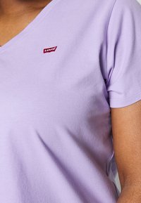 Liliowa bawełniana koszulka z dekoltem w serek, krótki rękaw oraz czerwoną metką Levi's po lewej stronie. Miękka faktura i gładkie wykończenie.