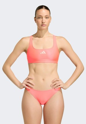 Kvinde stående med hænderne på hofterne iført koralfarvet sports-bh og matchende bikinitrusser mod en ensfarvet baggrund.