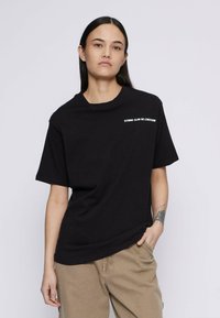 Persona ar gariem tumšiem matiem, ģērbta melnā T-kreklā ar baltu tekstu uz krūtīm un bēšās biksēs, pozē uz vienkārša gaiša fona.