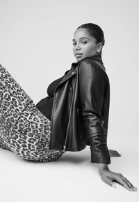 Veste de motard en cuir noir, haut noir et pantalon gris à imprimé léopard. Texture lisse ; modèle assis avec la main posée au sol, en noir et blanc.