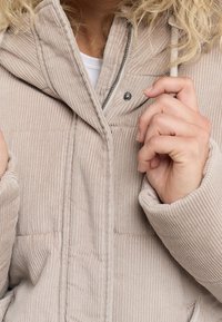 Beżowa kurtka z kordurowego materiału z kapturem, z zamkiem błyskawicznym i detalami na guziki. Tkanina ma pionowe prążkowanie i jest miękka w dotyku.