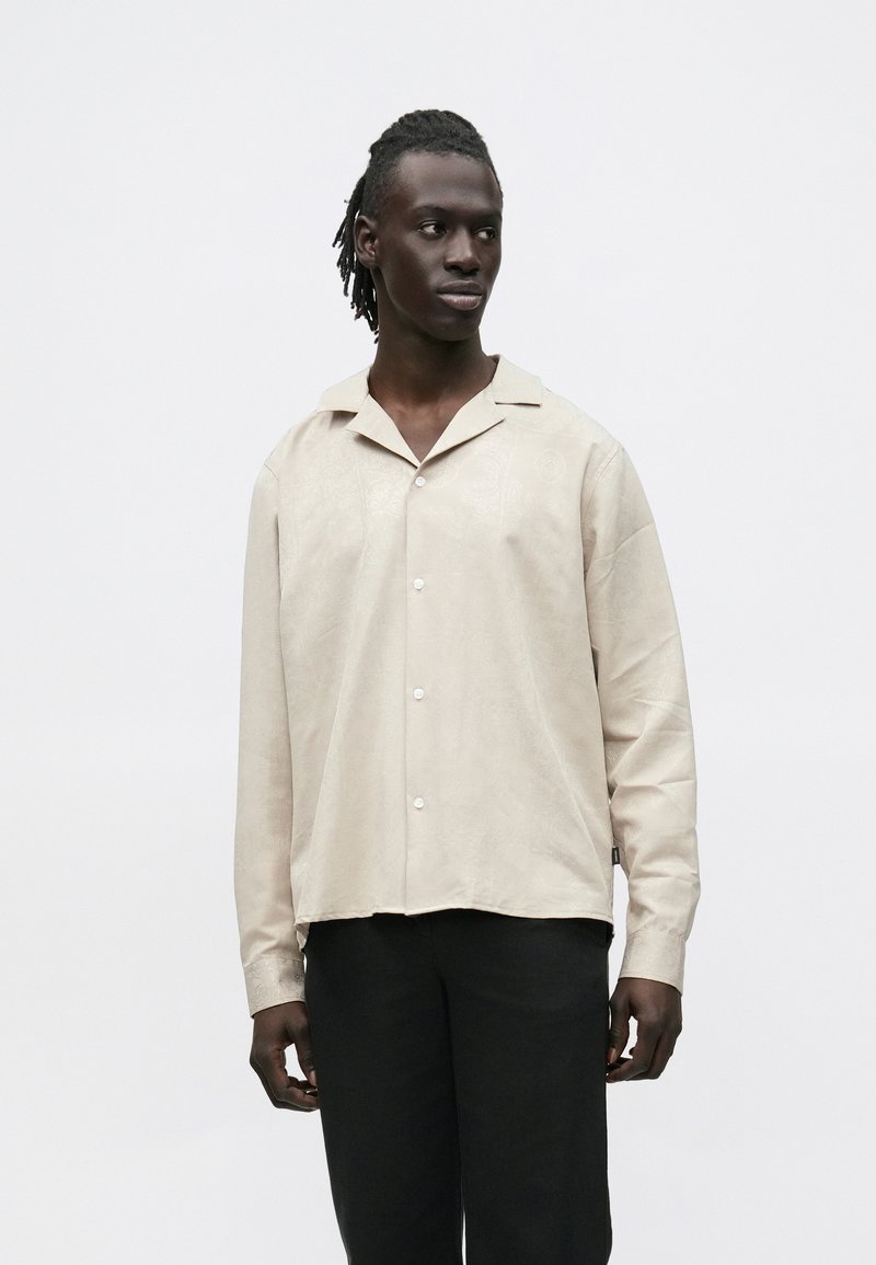 Jasnobeżowa koszula na guziki z wyciętym kołnierzem, długimi rękawami i subtelnym wzorem teksturowanym, w połączeniu z czarnymi spodniami.