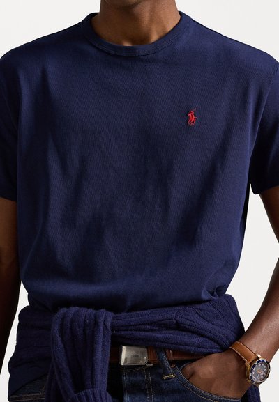 Sötétkék pamut póló kerek nyakkivágással és rövid ujjakkal, a mellkason piros hímzett logóval. Derékban megkötött sötétkék pulóverrel viselve.