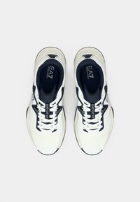 Fehér atlétikai cipő tengerészkék részletekkel, texturált hálós felsőrész, párnázott nyelv, valamint oldalán és talpbetétjén logórészlet.