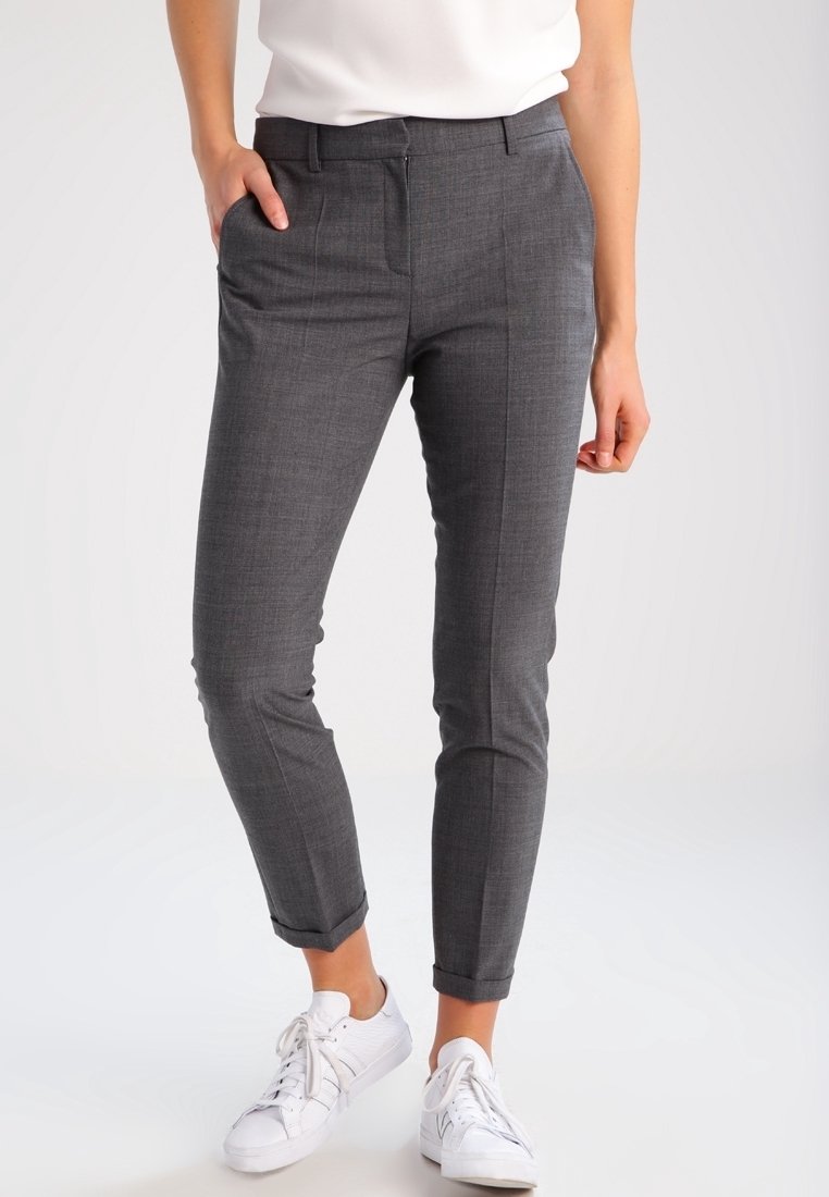 Personne portant un pantalon habillé gris slim avec des ourlets retroussés, associé à des baskets blanches et un haut blanc, debout avec une main dans la poche.