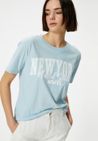Camiseta de algodón azul claro con texto blanco en negrita "NUEVA YORK Estados Unidos MMVII" en un corte relajado y cuello redondo clásico.