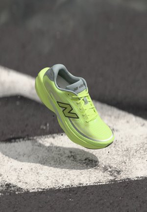 Φωσφοριζέ κίτρινο παπούτσι running της New Balance, αιωρούμενο πάνω από άσφαλτο με λευκές βαμμένες γραμμές και μικρά σκουπίδια από κάτω.