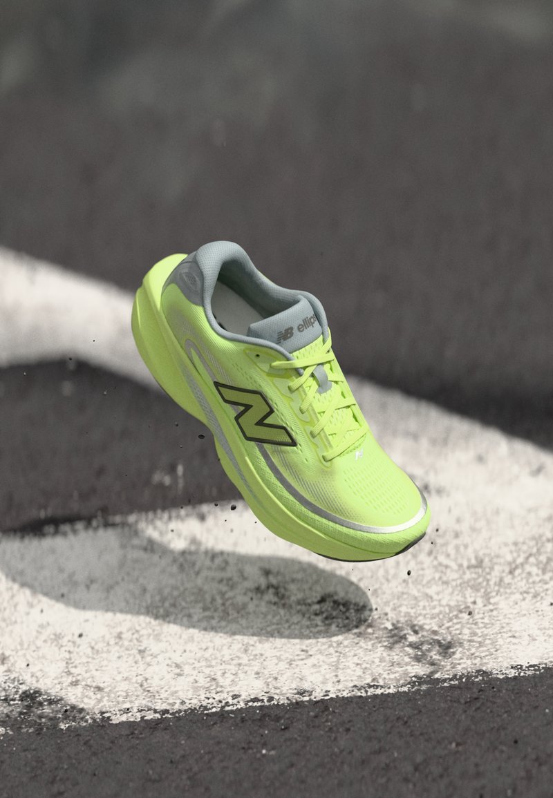 Φωσφοριζέ κίτρινο παπούτσι running της New Balance, αιωρούμενο πάνω από άσφαλτο με λευκές βαμμένες γραμμές και μικρά σκουπίδια από κάτω.