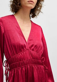 Fuchsia dress with a plunging V-neck, elasticized waist, and drawstring details. The fabric has a crinkled texture and long sleeves.