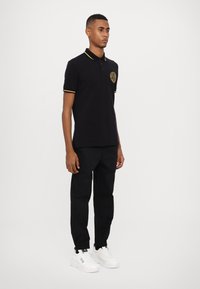 Tricou polo negru cu detalii galbene și logo, purtat cu pantaloni negri și adidași albi, evidențiind un stil sportiv clasic.