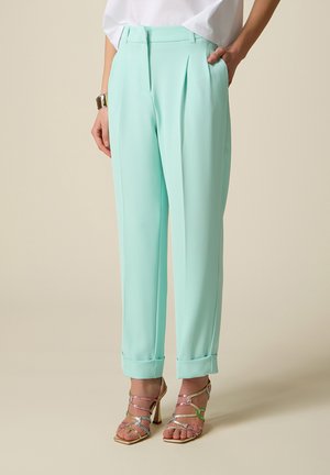 Kobieta ubrana w miętowo-zielone spodnie z wysokim stanem i podwiniętymi nogawkami, zestawione z wielokolorowymi sandałami na wysokim obcasie z paskami oraz białą, włożoną do środka bluzką.