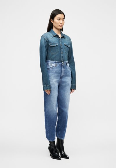 Γυναίκα που στέκεται φορώντας σκούρο πουκάμισο από τζιν, ανοιχτόχρωμο τζιν με ψηλή μέση και μαύρα μυτερά μπότες με τακούνι σε λευκό φόντο.