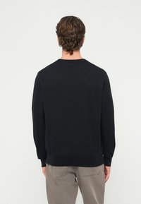 Άνδρας με καστανά σγουρά μαλλιά, φοράει μαύρο πουκάμισο με μακριά μανίκια και γκρι παντελόνι, στέκεται με την πλάτη προς την κάμερα μπροστά σε λευκό φόντο.