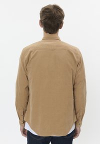 Lichtbruine langslavige shirt met een gladde textuur, met knoopmanchetten en een afgeronde zoom, op een eenvoudige achtergrond.