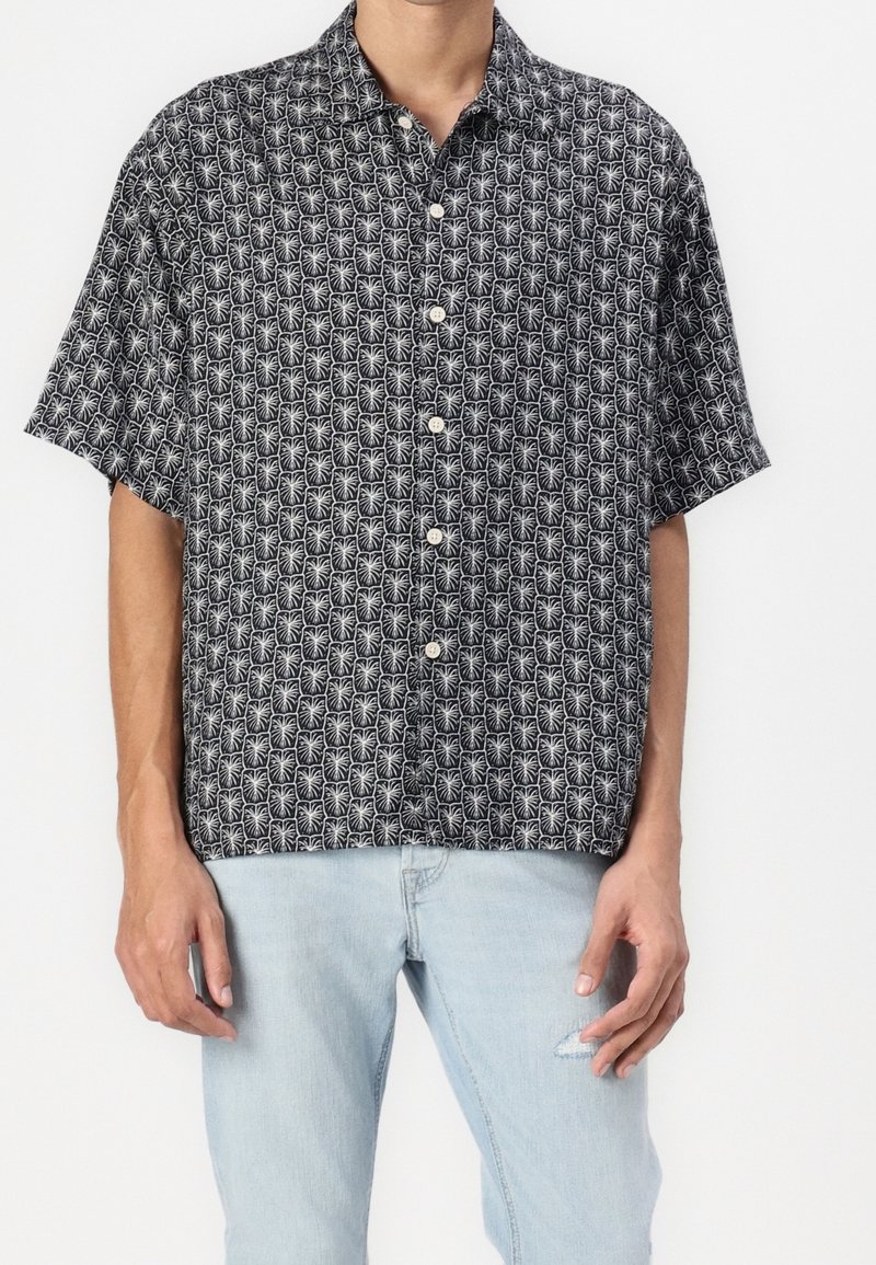 Homme portant une chemise noire à manches courtes avec des motifs de palmiers blancs et un jean bleu clair déchiré, debout sur fond blanc.