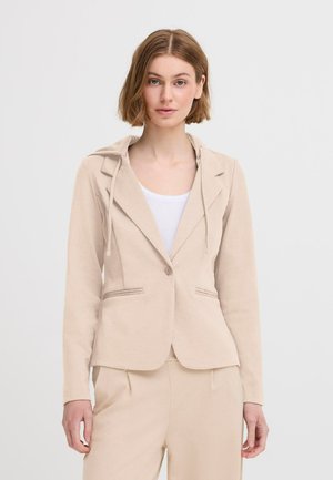 Beige taillierter Blazer mit Kapuze, Einknopfverschluss und Vorderentaschen. Texturiert mit glatter Oberfläche. Kombiniert mit einem weißen Top.