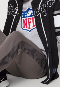 Persona che indossa una maglietta grigia con il logo NFL, una giacca nera e bianca dei Chicago, pantaloni grigi a fantasia e sneakers nere della Nike, in una posa seduta.