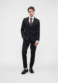 Jovem em fato preto com camisa branca, gravata bordô e sapatos pretos a vestir, de pé contra um fundo branco simples.