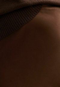 Een bruine geribbelde gebreide kraag valt over een gladde, donkerbruine stof, waardoor een textuurcontrast ontstaat in een close-up van een kledingstuk.