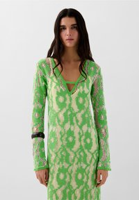 Brede gebreide jurk met een diepe V-hals, lange mouwen, in helder groen met een gestructureerd bloemenpatroon en lichte beige accenten. Zwarte armband om de pols.