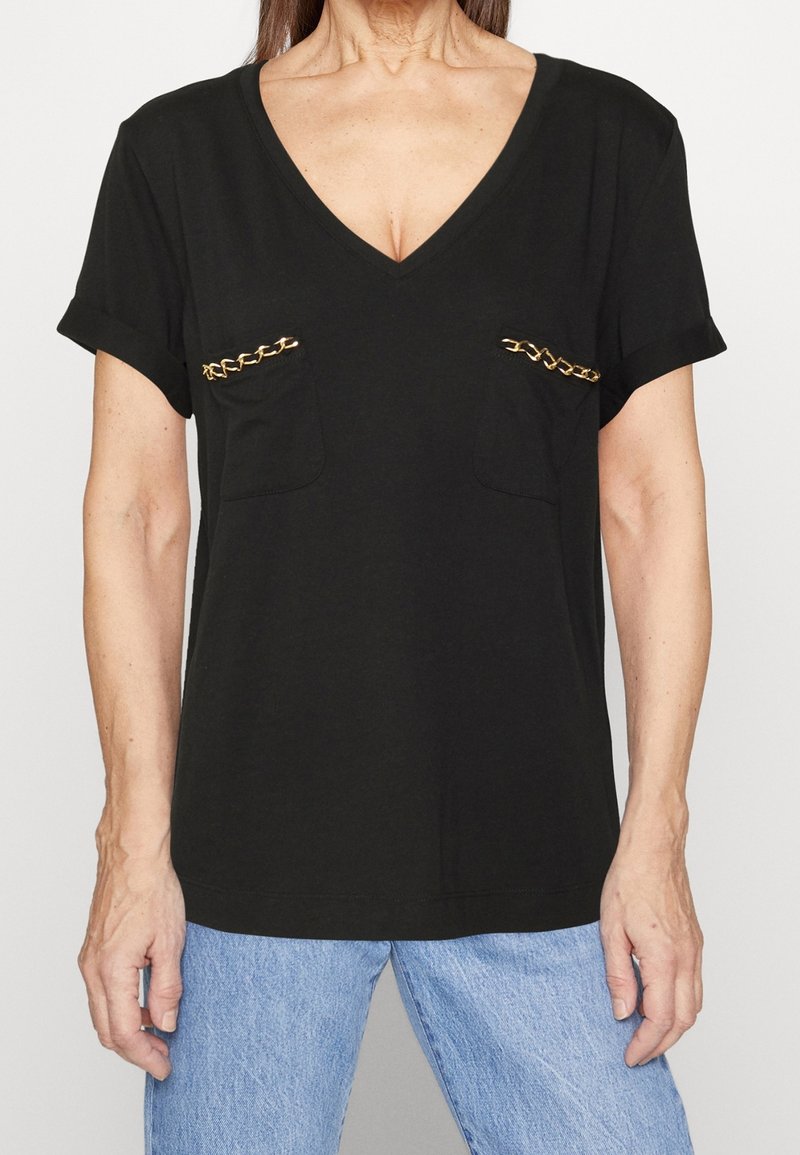 Camiseta negra de cuello en V con mangas enrolladas, que cuenta con dos bolsillos frontales adornados con detalles de cadena dorada. Tela suave, ajuste relajado.