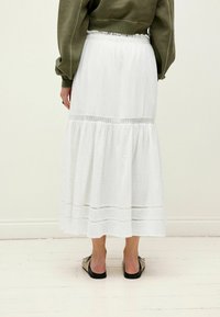 Jupe maxi blanche, à niveaux, fabriquée en tissu texturé avec des accents de dentelle ; associée à un haut gris-vert et des sandales plates à motifs.