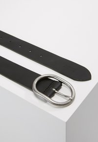 Ceinture en cuir noir avec une boucle ronde en métal argenté, présentant une surface lisse et plusieurs trous d'ajustement le long de la sangle.
