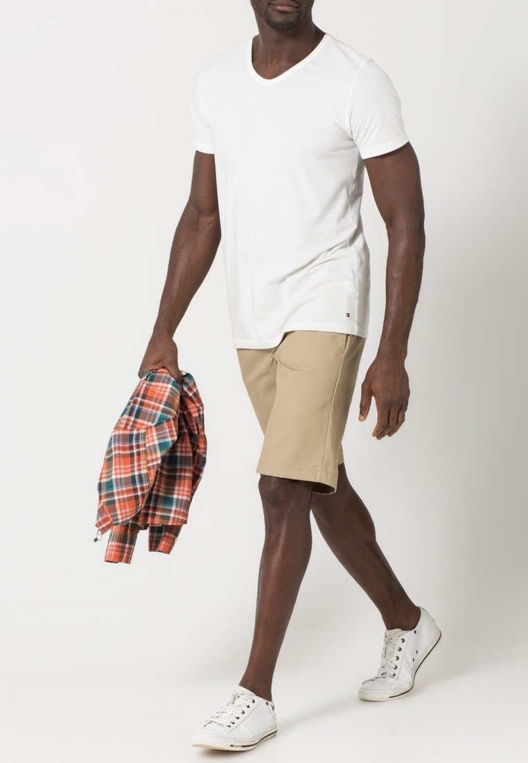 Vit t-shirt med v-ringad hals i kombination med beige shorts. Personen håller en röd och grön rutiga jacka och har vita sneakers på sig.