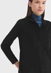 Čierna manžetová košeľa s dlhými rukávmi, gombíkovým zapínaním, náprsné vrecko a golier; jemná textúra a jemné ryhy viditeľné v látke.