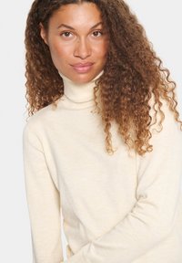 Light beige turtleneck sweater in a soft, ribbed texture. Features long sleeves and a fitted silhouette. Background is plain gray.