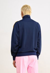 Marinblå sportjacka med hög krage, långa ärmar och gula sidopaneler, tillverkad av ett mjukt, andningsbart material.