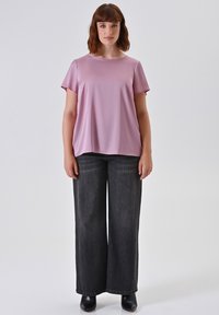 Frau steht, trägt ein lockeres, rosa T-Shirt mit kurzen Ärmeln, weite schwarze Jeans und schwarze Stiefeletten vor einem schlichten, hellen Hintergrund.