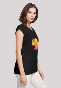 Schwarzes Kurzarm-T-Shirt mit einem bunten, cartoonhaften gelben Triceratops-Graphic auf der Vorderseite. Weiße Hose sichtbar.