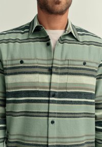Man wearing green, beige, and black striped button-up shirt over a light beige undershirt, with visible chest and neck area.