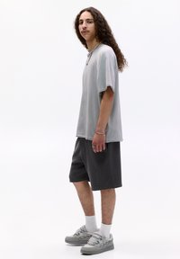Šedé oversized tričko s krátkým rukávem, spárované s tmavě šedými šortkami. Model má na sobě světle šedé tenisky a bílé ponožky, doplněné náhrdelníkem a prsteny.