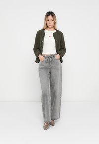 Olivenfarget, strukturert cardigan over en hvit, kort skjorte med røde hjerter. Lys grå, vide jeans og taupe hæler fullfører antrekket.