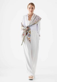 Chemise blanche à boutons, superposée avec une écharpe à motifs, comprenant des designs floraux et géométriques, associée à un pantalon large blanc et des sandales.