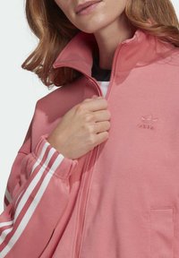 Giacca rosa con zip e colletto alto, strisce bianche di accento sulle maniche e logo ricamato sul petto. Materiale morbido e liscio con vestibilità rilassata.