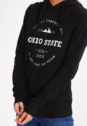 Osoba ubrana w czarną bluzę z kapturem, z nadrukiem białym na przodzie: „Ohio State”, „Ft Campbell Hill 1995” oraz „We are living our dream”.