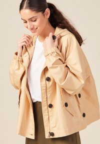 Giacca beige con cappuccio, polsini elasticizzati, chiusura frontale con zip e grandi bottoni marroni. Tessuto leggero con un design rilassato e squadrato e tasche laterali.