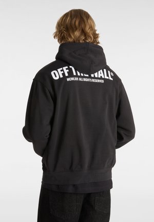 Personne aux cheveux mi-longs portant un sweat à capuche noir avec le texte « OFF THE WALL MCMLXVI ALL RIGHTS RESERVED » dans le dos sur un fond uni.